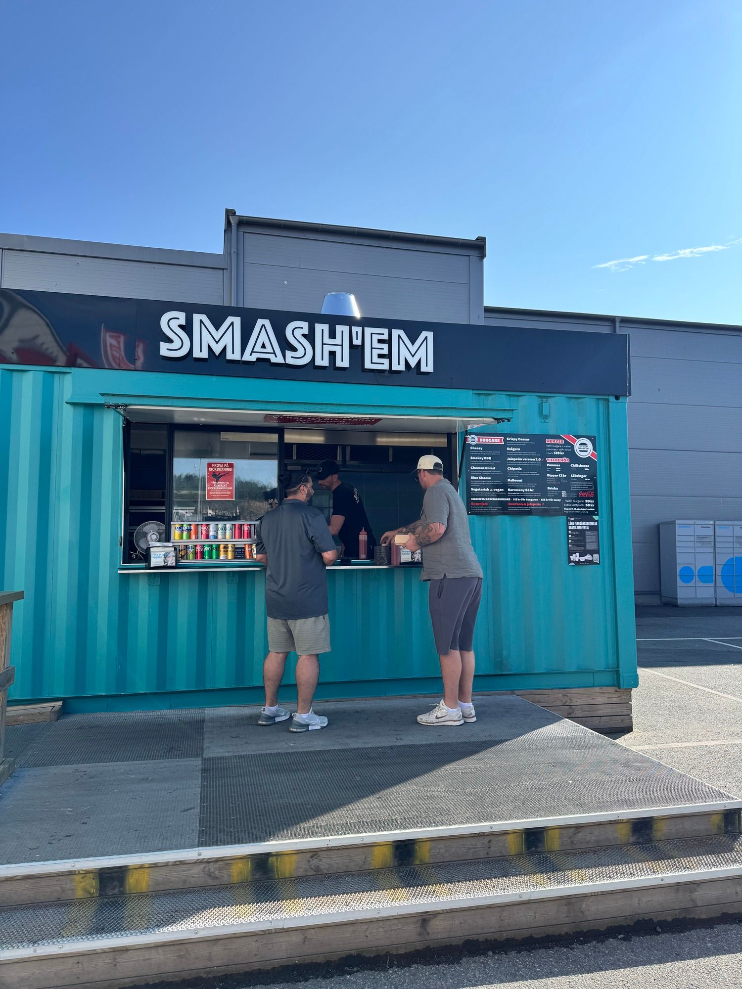Två gäster står vid Smash'ems öppna serveringslucka i den turkosa containern på Bultgatan 35 i Kungälv, med trädäck och trappa framför.